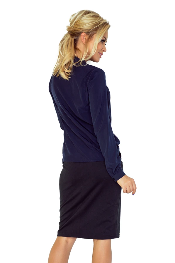 Sieviete ir ģērbusies tumši zilā blūzē ar elegantu sasienamu detaļu priekšpusē. Blūze ir kombinēta ar melnu svārku, radot stilīgu un profesionālu izskatu.
