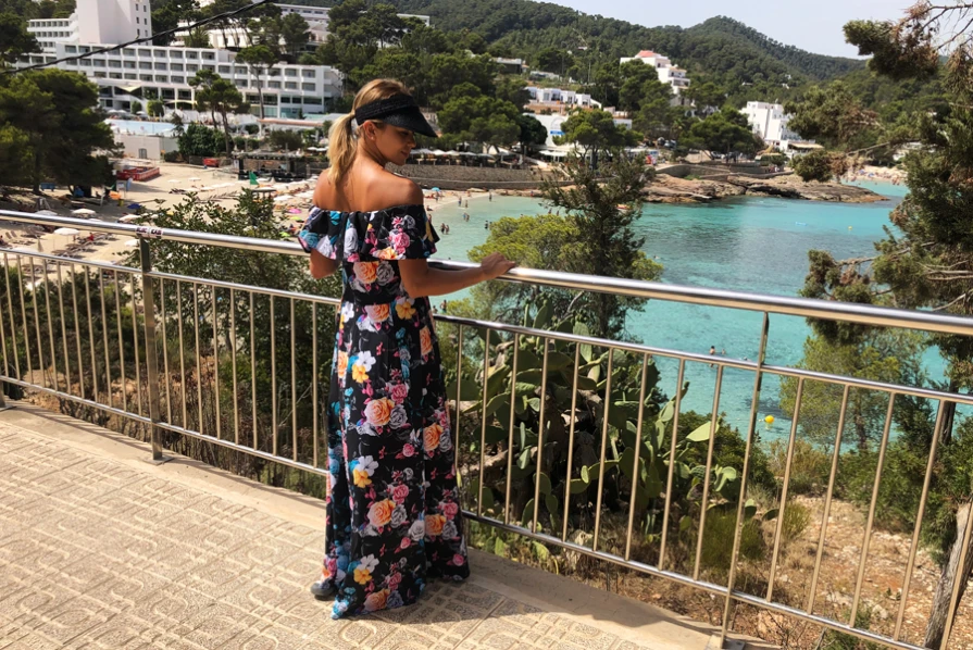 Uz fotoattēla redzama sieviete, kas valkā garu, ziedu raksta kleitu ar spāņu stila plecu izgriezumu. Kleita ir koša un eleganta, ideāli piemērota vasaras pastaigām pie jūras.