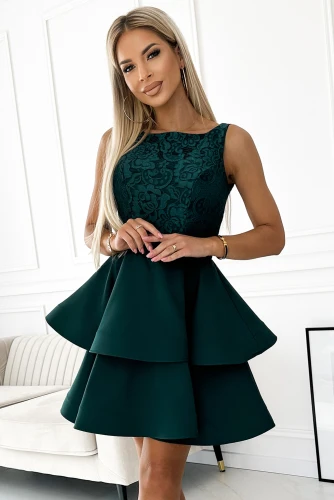 Sieviete valkā elegantu, tumši zaļu kleitu ar divkāršu svārku daļu un mežģīņu augšdaļu. Kleita ir piemērota svinīgiem pasākumiem, izceļot sievišķību un stilu.}